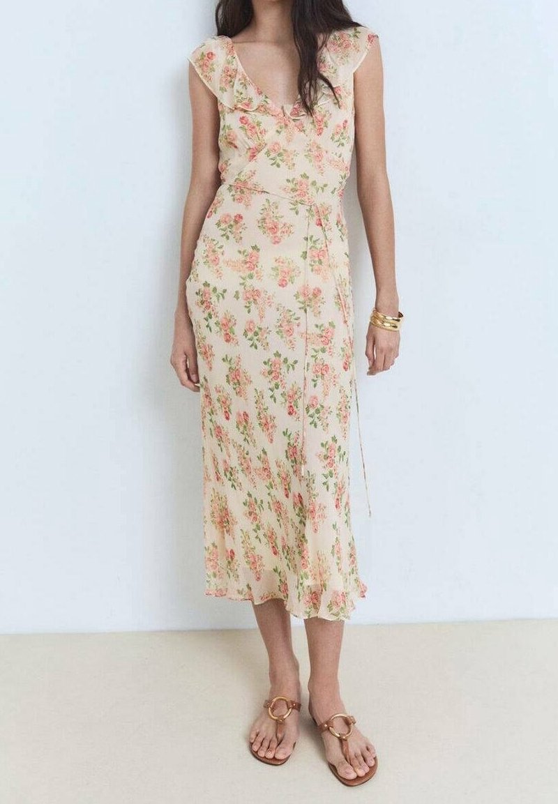 Robe midi fleurie de couleur pêche pâle avec des manches à volants, arborant des fleurs vertes et roses. Portée avec des sandales plates marron. Texture de tissu lisse.