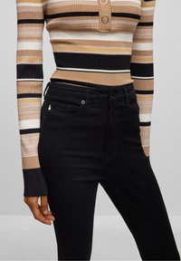 Top a rayas con textura acanalada en beige, negro y blanco, con escote abotonado, combinado con jeans negros de diseño ajustado y detalles sutiles.