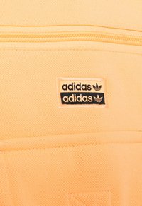 Close-up van lichtoranje stof met een zwart-oranje Adidas-logo patch bij een ritssluitingnaad.