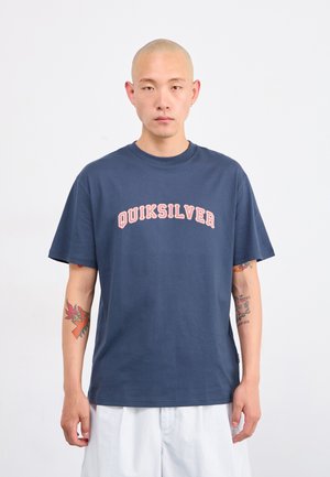 Mann med tatoveringer iført en marineblå Quiksilver t-skjorte og hvite shorts mot en enkel hvit bakgrunn, ser fremover.
