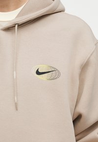 Μπεζ φούτερ με κουκούλα, με μαύρο λογότυπο Nike μέσα σε οβάλ σχήμα με κίτρινο και γκρι σχέδιο πλέγματος στο στήθος, διαθέτοντας ρυθμιζόμενο κορδόνι στην κουκούλα.