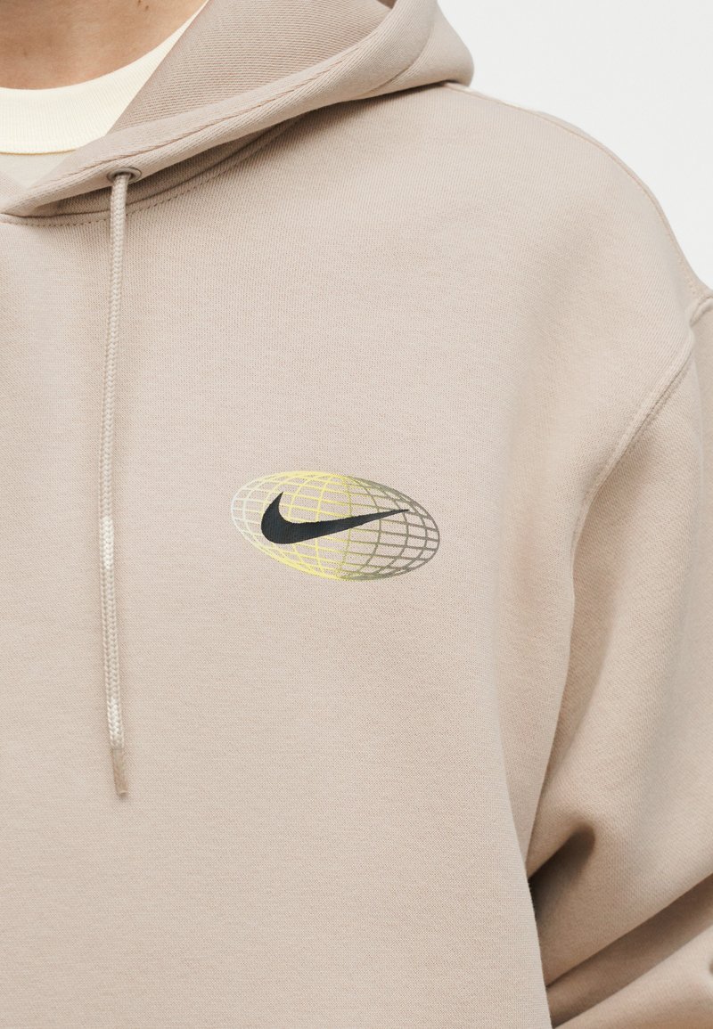 Felpa beige con cappuccio caratterizzato da cordoncino, con un swoosh Nike nero all'interno di un ovale a motivo a griglia giallo e grigio sul petto.