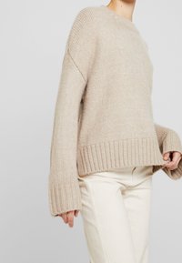 Pull en tricot beige avec un col rond, des poignets et un ourlet côtelés. Il présente un tissu texturé et une coupe décontractée, porté sur un pantalon blanc.