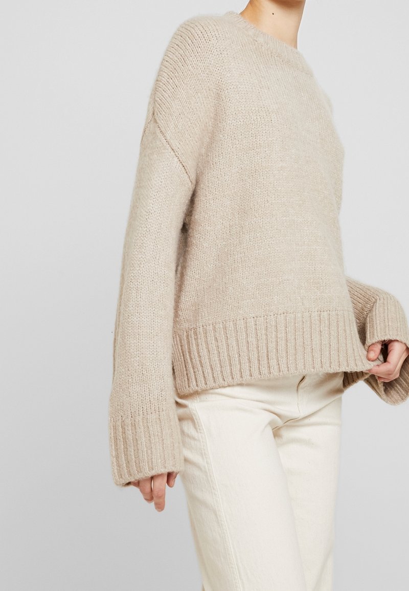 Pull en tricot beige avec un col rond, des poignets et un ourlet côtelés. Il présente un tissu texturé et une coupe décontractée, porté sur un pantalon blanc.