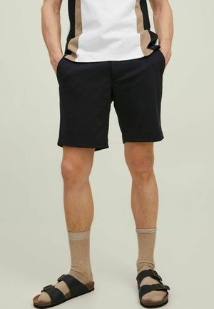 Person iført sorte shorts, beige sokker, sorte sandaler med spænde og en hvid og beige color-blocked top med hænderne i lommerne.