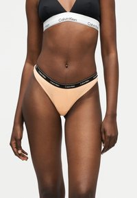 Γυναίκα που φοράει μαύρο και άσπρο σουτιέν Calvin Klein και ροζιές πυτζάμες με μαύρη ζώνη Calvin Klein σε απλό φόντο.