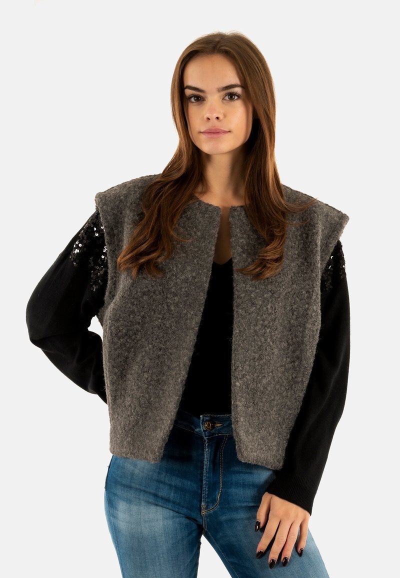 Gilet gris en feutre avec un design court, doté de manches en sequins noirs et d'un devant ouvert. Porté sur un haut noir et un jean en denim bleu.