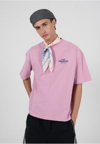 Roosa lühikeste varrukatega puuvillast t-särk, millel on tumesinise niidiga tikitud sõna "KEELATUD". Kandmise juurde kuuluvad heledavärviline mustriline sall ja hall müts.