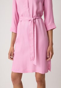 Abito rosa con cintura, chiusura a bottoni, maniche a tre quarti e spacchi laterali. Realizzato in tessuto morbido con una leggera lucentezza.