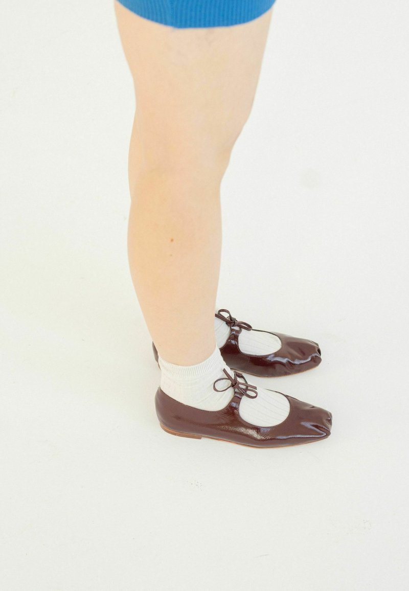 Bailarinas de charol marrón con punta redondeada, que presentan un diseño recortado y lazos, combinadas con calcetines blancos hasta el tobillo. Textura suave.