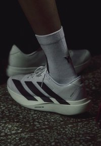 Personne portant des chaussures de course Adidas blanches avec des rayures noires et des chaussettes blanches sur un sol en terrazzo sous une lumière faible.