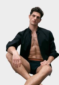 Camisa negra de botones, parcialmente abierta, sobre ropa interior negra ajustada. Tela suave, diseño a medida, con un detalle en la cintura grabado.