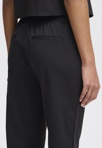 Black tapered trousers with an elastic waistband, smooth texture, two back pockets, and a subtle sheen.