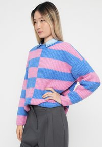 Ženska z dolgimi blond lasmi, oblečena v pulover z roza in modrimi črtami, čez svetlo modro srajco s ovratnikom in sivo hlačami.