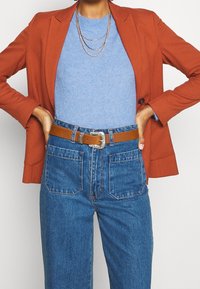 Roestkleurig colbert over een lichtblauwe trui, gecombineerd met een high-waisted blauwe jeans. Bruine riem met een zilveren gesp accentueert de outfit.