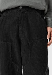 Pantaloni in corduroy neri con evidenti righe verticali, due grandi tasche anteriori e vita aderente con un singolo bottone in metallo.