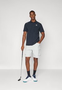 Rövid ujjú, sötétkék golfpóló, zsebbel és logóval, világosszürke cargo rövidnadrággal párosítva. A modellen golfütő van, fehér cipőt visel kék részletekkel.