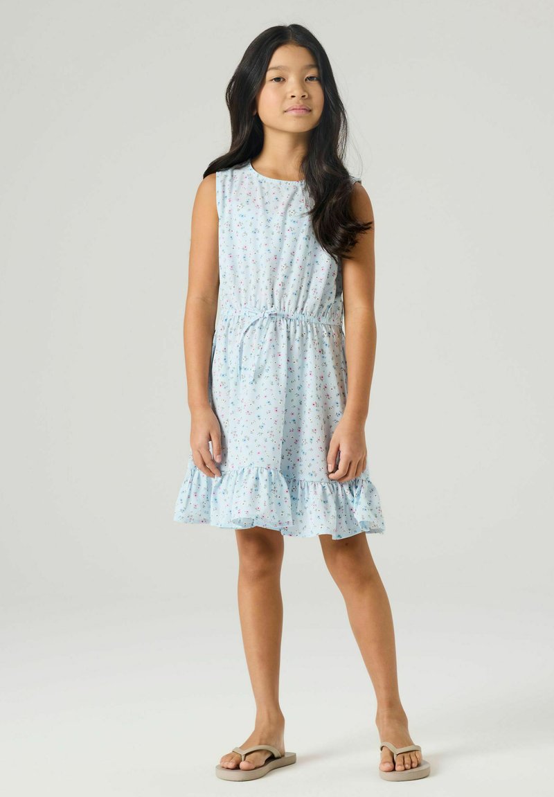 Fille aux longs cheveux noirs portant une robe sans manches bleu clair avec un petit imprimé floral et un ourlet à volants, debout en tongs beiges.