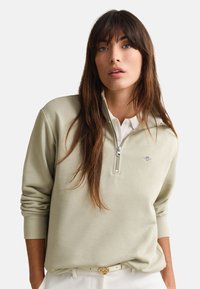 Γυναίκα με μακριά καστανά μαλλιά που φοράει ένα μπεζ φούτερ με φερμουάρ μέχρι τη μέση πάνω από ένα λευκό πουκάμισο με γιακά, ποζάροντας με τα χέρια στις τσέπες μπροστά σε λευκό φόντο.