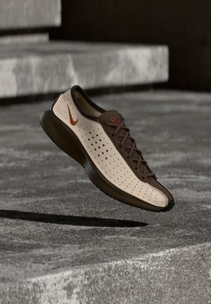 Nike-sneaker met een beige geperforeerde bovenkant en bruine accenten zweeft boven betonnen trappen en werpt een schaduw.