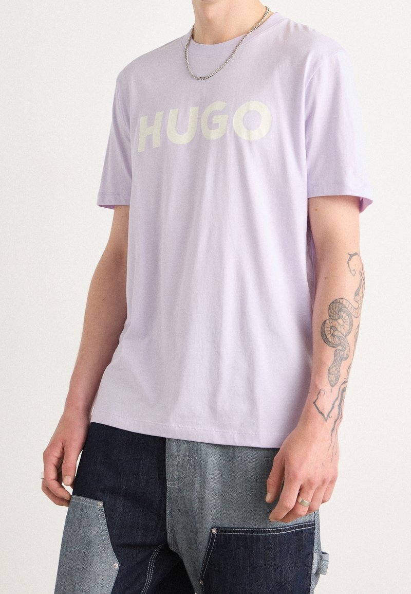 T-shirt en coton lavande présentant un grand logo blanc "HUGO", encolure ras du cou, manches courtes et coupe décontractée. Porté avec un pantalon en denim patchwork.