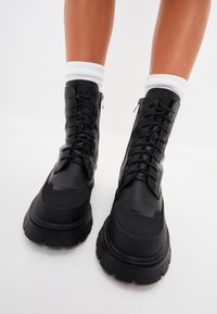 Botas de combate de cuero negro con una suela gruesa, que cuentan con cordones, cremalleras laterales y acentos texturizados en la zona de los dedos. Calcetines blancos visibles por encima.