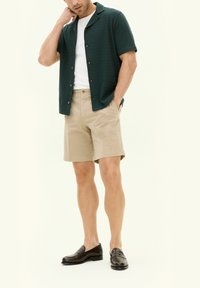 T-paita, jossa lyhyet hihat ja tummanvihreä väritys, kuvioitu kangas, valkoinen t-paita, beige lyhyet housut ja mustat loaferit. Malli seisoo käsi niskassa.