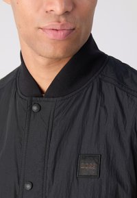 Schwarze Bomberjacke aus leichtem Material, mit einem gerippten Kragen, Knopfverschluss und einem quadratischen Leder-Logo-Patch.