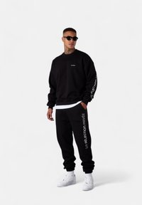 Man wearing black sweatshirt and sweatpants with white text, white sneakers, and sunglasses, standing against a plain white background.