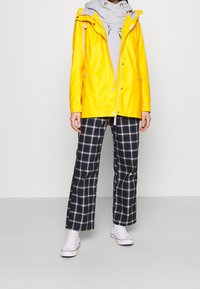 Gul vattentät jacka med huva, grå sweatshirt under, och bredrandiga byxor i svart och vitt. Vita sneakers kompletterar outfiten.