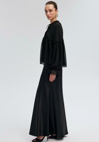 Black long-sleeve dress with a high neck, sheer fabric, and loose fit. Features gathered accents and bell sleeves, worn with black heeled sandals.