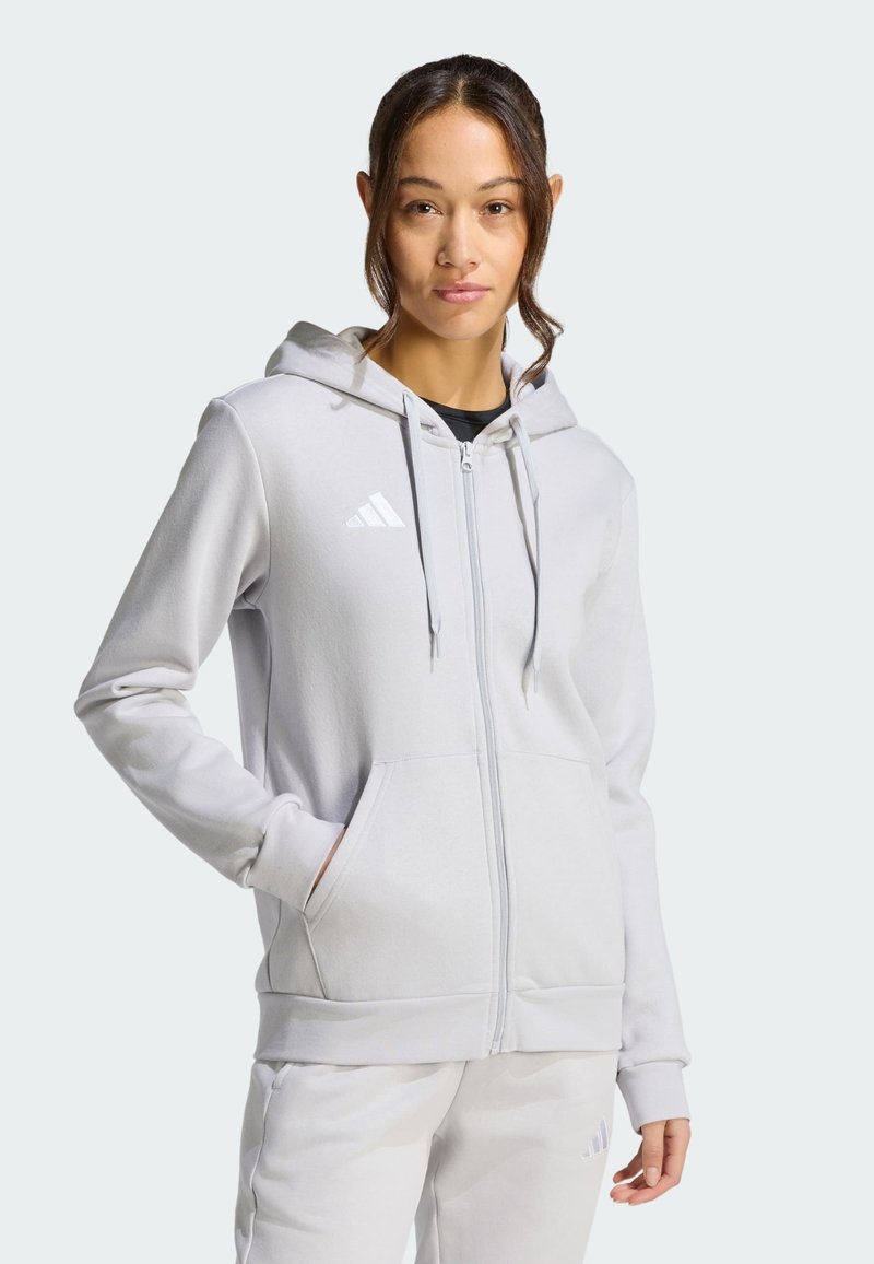 Femme portant un sweat à capuche zippé Adidas gris clair et un pantalon de survêtement assorti, debout avec une main dans la poche devant un fond uni.