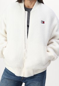 Persoon in een witte sherpa ritsjack met het Tommy Jeans-logo op de borst, grijze shirt eronder en blauwe spijkerbroek, met handen in de zakken.