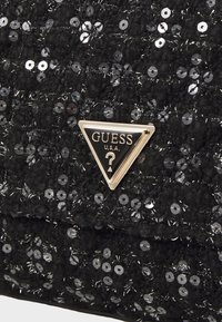 Czarna torebka wykonana z teksturowanej tkaniny, zdobiona srebrnymi cekinami. Na przodzie znajduje się trójkątne logo Guess w kolorze złotym i czarnym.