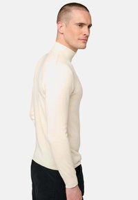 Homme avec les cheveux courts portant un pull-over en col roulé beige ajusté et un pantalon noir, se tenant de profil contre un fond uni.