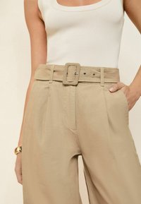 Beige high-waisted broek met een wijde pijpdesign, met een vierkante riemgesp en plooien, gecombineerd met een witte geribbelde tanktop.