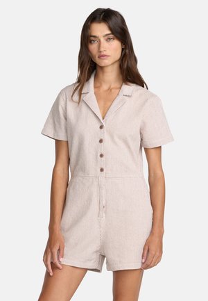 Kobieta w beżowo-białym, paskowanym, krótkim kombinezonie z kołnierzykiem i guzikami, stojąca na jednolitym jasnym tle.