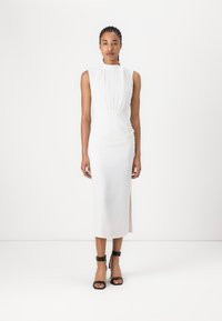 White sleeveless dress with a high neckline, gathered waist, and side slit. Black ankle-strap sandals complement the look.