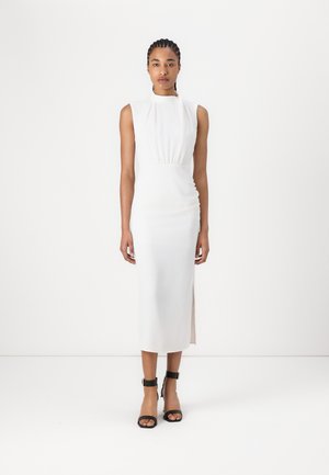 White sleeveless dress with a high neckline, gathered waist, and side slit. Black ankle-strap sandals complement the look.