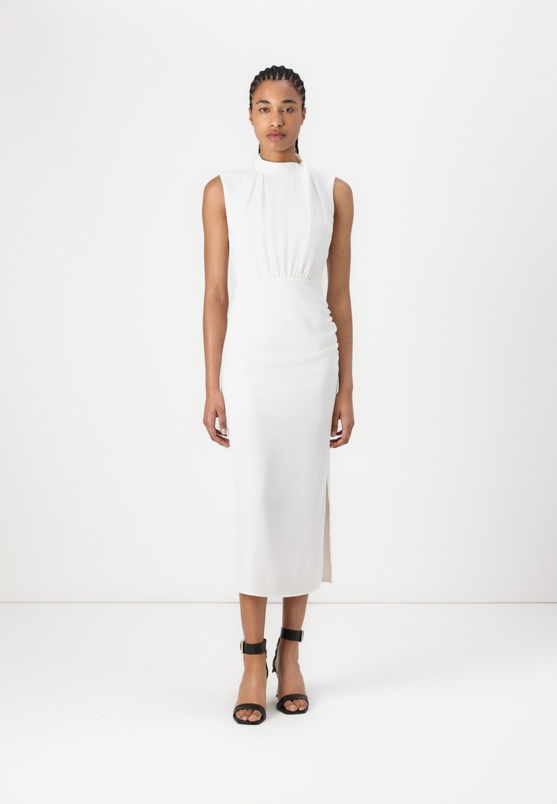 White sleeveless dress with a high neckline, gathered waist, and side slit. Black ankle-strap sandals complement the look.