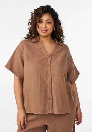 Naine lokkis juustega, seljas lahtine pruun lühikeste varrukatega nööpidega särk ja sobivad püksid, seisab valge tausta ees.