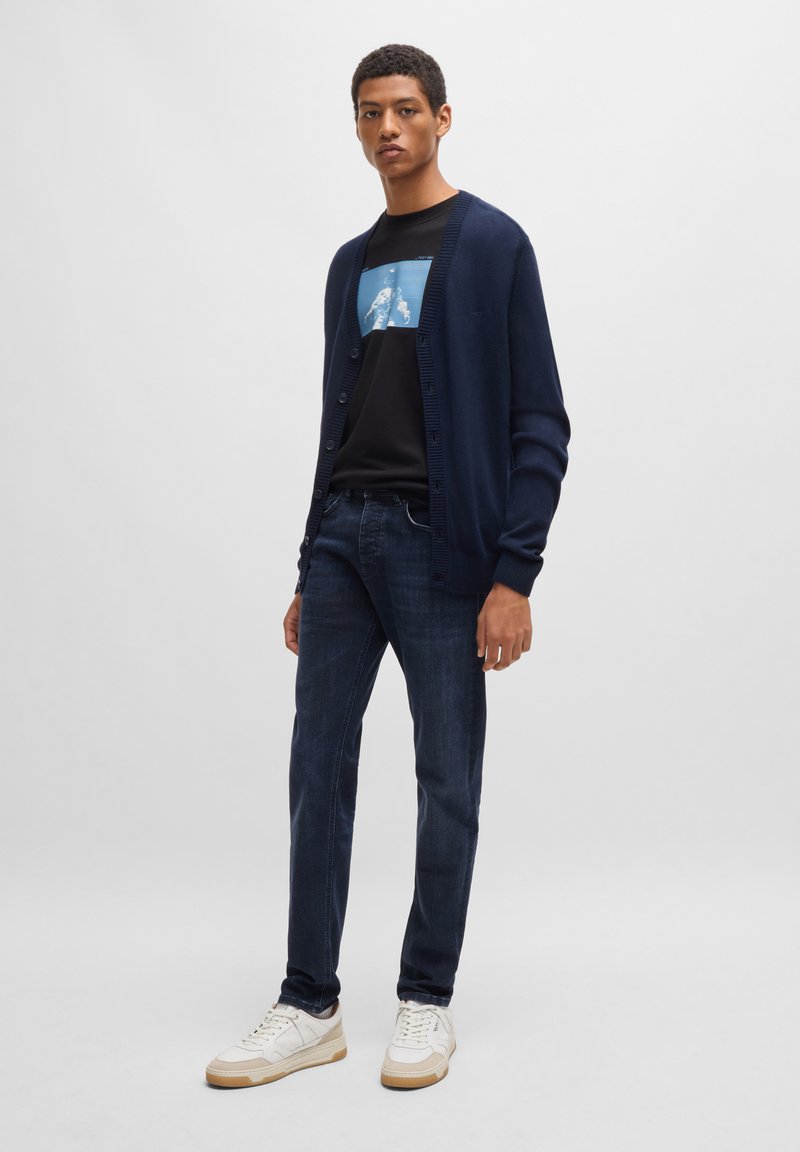 Marineblauer Strickcardigan, schwarzes Grafik-T-Shirt, dunkelblaue Jeans und weiße Sneaker. Einfaches Design mit Knöpfen und gerippten Bündchen.