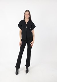 Schwarzes kurzärmliges Oberteil mit tiefem V-Ausschnitt und einem auffälligen vorderen Knopf. Kombiniert mit schwarzen ausgestellten Hosen und schwarzen Stiefeln mit spitzem Fuß.