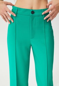 Personne portant un pantalon taille haute vert vif avec poches latérales, bouton et fermeture éclair, les mains tirant légèrement sur la ceinture.