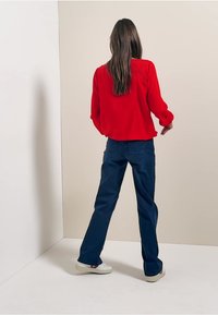 Blusa rossa a maniche lunghe con vestibilità ampia, abbinata a pantaloni larghi blu scuro e sneakers bianche, su uno sfondo neutro.