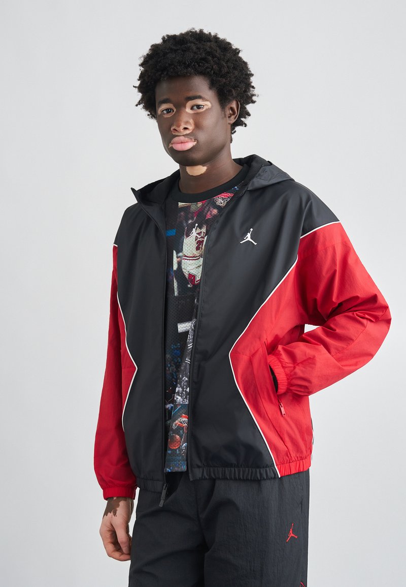 Μαύρο και κόκκινο αδιάβροχο μπουφάν με κουκούλα, με λευκές λεπτομέρειες, ελαστική μέση και λογότυπο στο στήθος. Ελαφρύ, με λεία υφή ύφασμα.