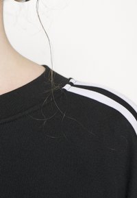 Pull noir côtelé avec un col rond et des accents rayés blancs sur les épaules ; la texture est légèrement surélevée et ajustée.