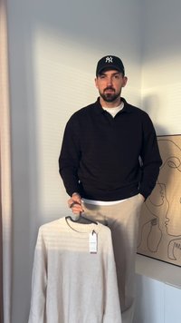 Schwarzes Langarm-Polohemd, weißes Unterhemd, beigen Hosen. Hält einen cremefarbenen Pullover mit Rundhalsausschnitt und Preisschild.
