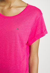 Camiseta de manga corta de color rosa brillante con una textura suave y mangas enrolladas, que presenta un pequeño logo en el pecho.