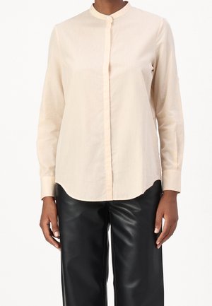 Personne portant une chemise beige à manches longues boutonnée avec un col mandarin et un pantalon en cuir noir, debout les mains détendues sur les côtés.
